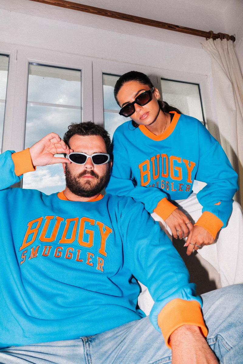 Man and Women wearing blue crewneck jumper with orange writing on front, in a lounge room 