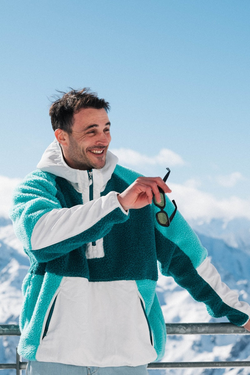 Man in a teal and white jacket holding sunglasses with snow-covered mountains in the background