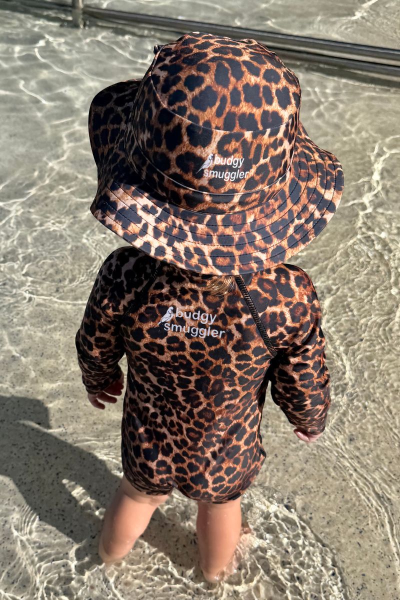 Child wearing a leopard print swimsuit and hat with 'Budgy Smuggler' branding, standing in shallow water.