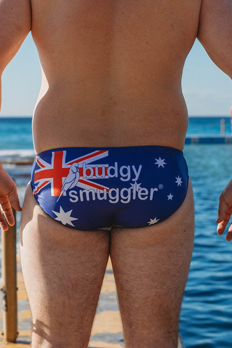 Rear view of a person wearing swim briefs with an Australian flag design and the text 'buddy smuggler' printed across the back, standing outdoors near water.