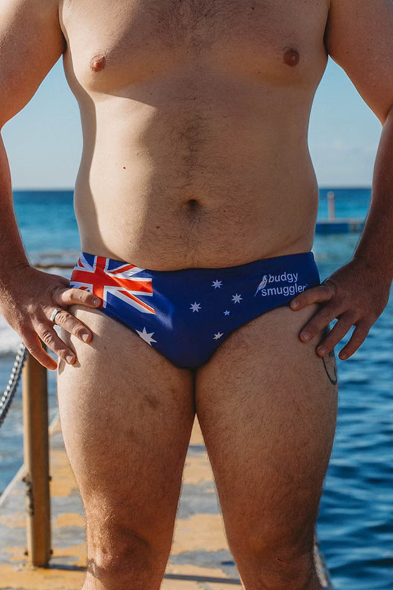 A close-up lifestyle shot of a man wearing blue swim briefs featuring the Australian flag and the white text 'budgy smuggler' on the side, standing on a wooden dock with the ocean in the background.
