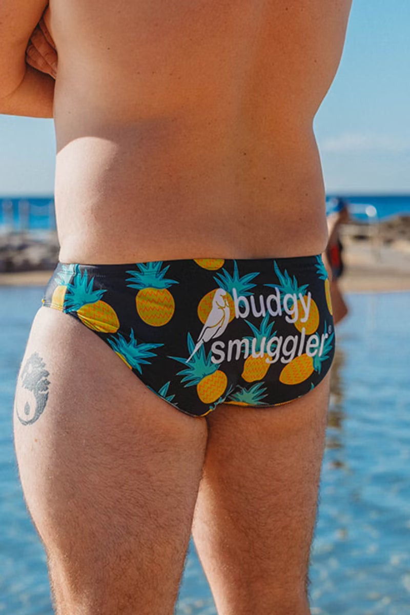 Rear view of a man wearing swim briefs with a black background, yellow pineapple print, and a white 'buddy smuggler' logo, standing in shallow blue water outdoors.