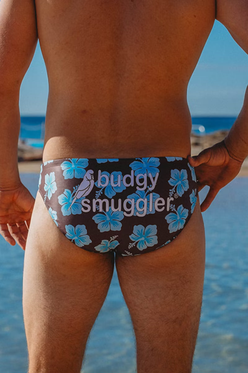 Rear view of a man standing on a beach wearing dark brown swim briefs with a blue hibiscus floral pattern and the text 'buddy smuggler' printed across the back.