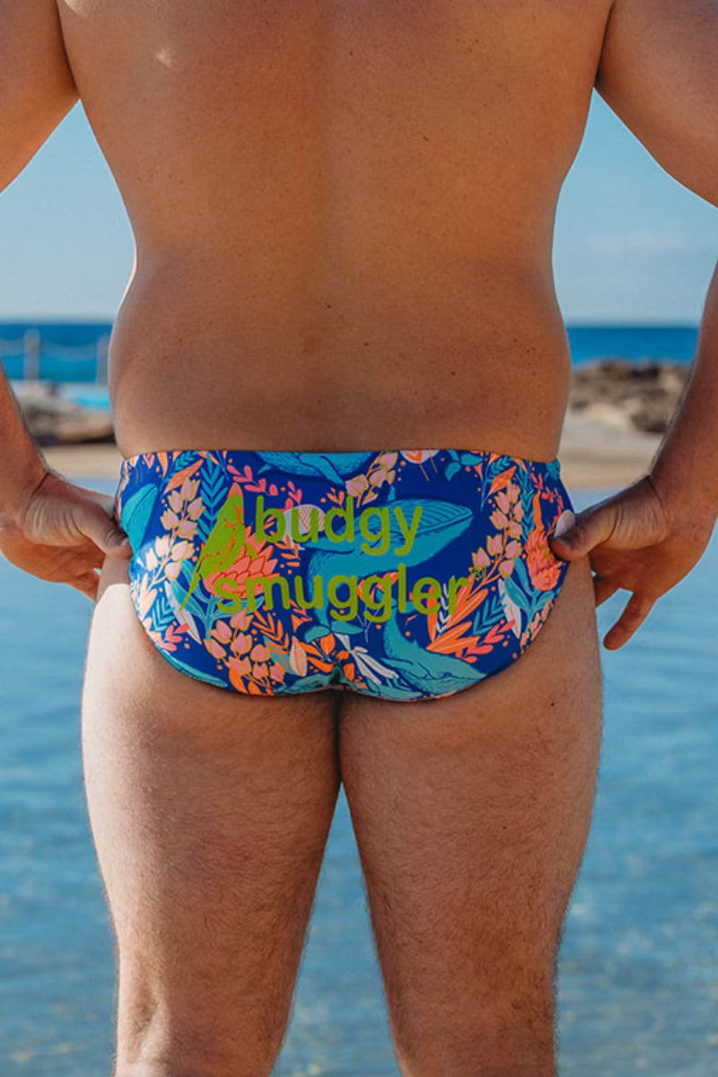 Rear view of a man standing at the beach wearing low-rise swim briefs with a blue tropical leaf pattern and the text 'Happy Snuggler' printed in yellow-green.