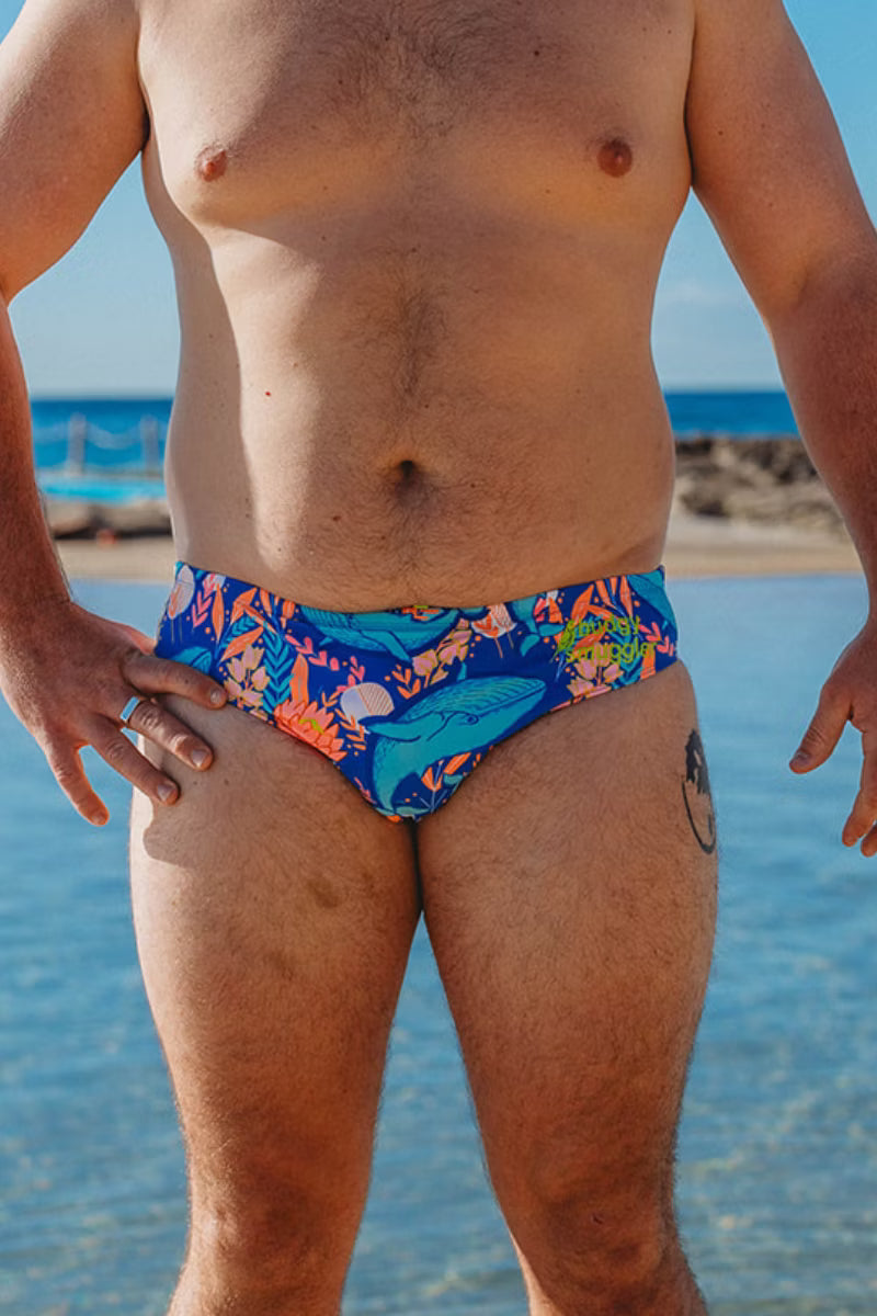 Front view of man in Blue Whale swimmers, a royal blue whale and tropical leaf print, by the ocean