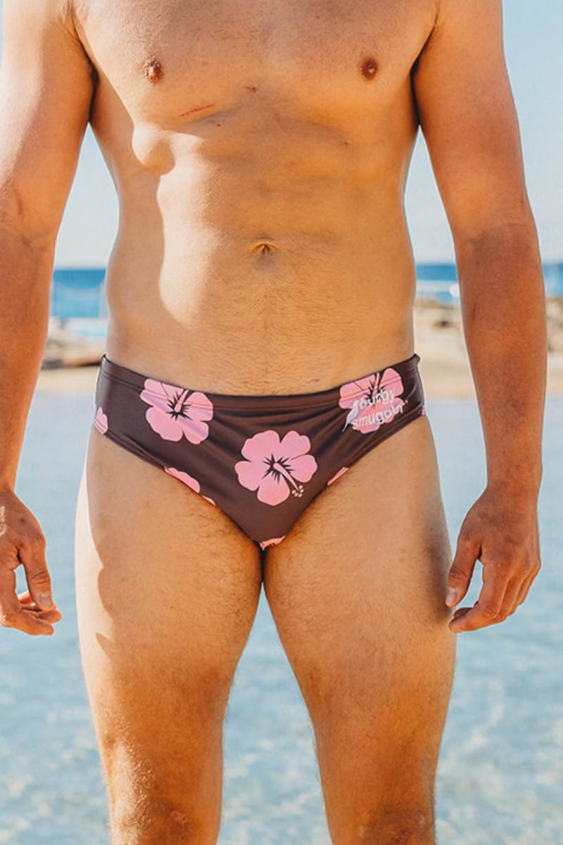A close-up of a man's lower torso and thighs wearing low-rise swim briefs with a dark brown base and pink hibiscus floral print, set against a sunny beach background.