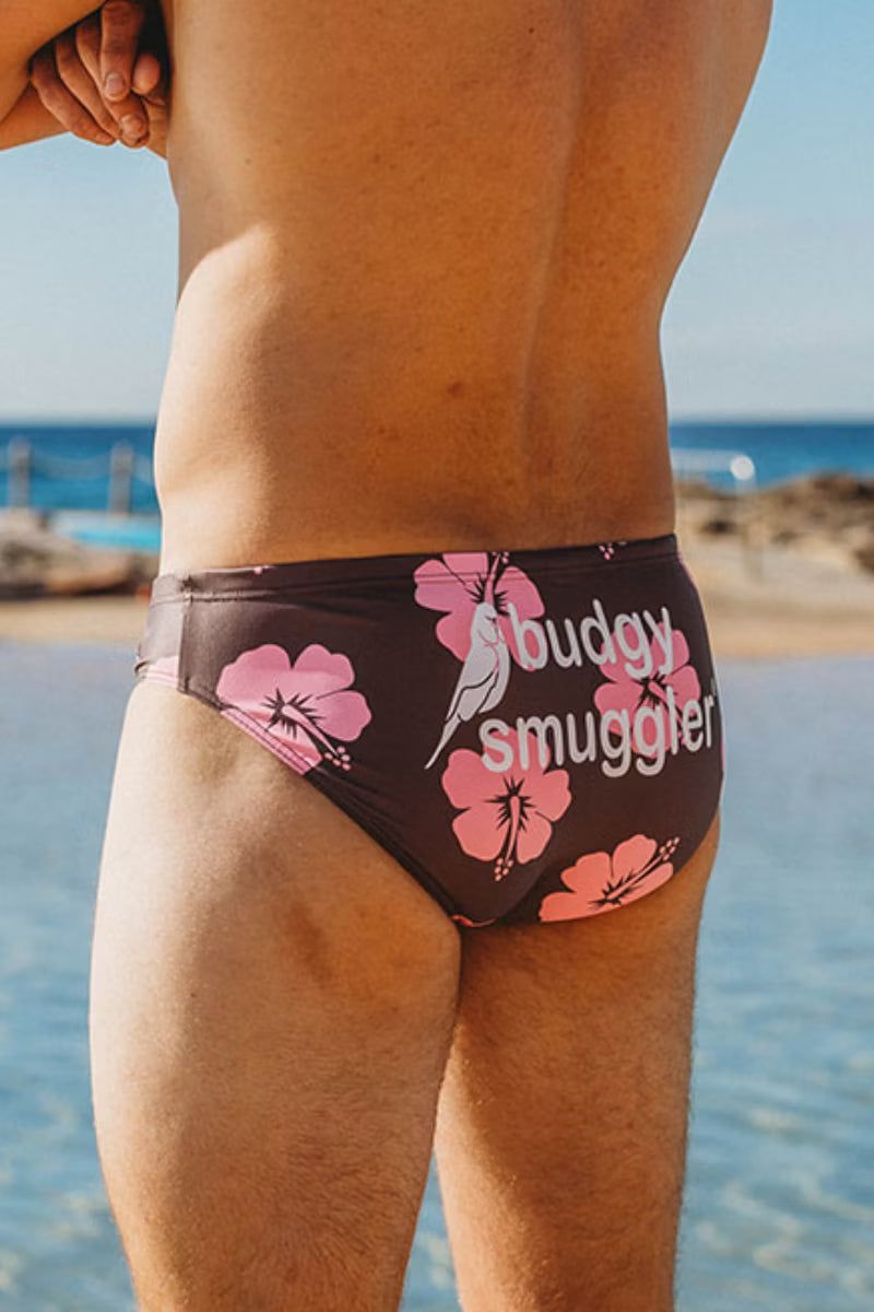 A close-up back view of a man wearing bikini bottoms at the beach. The swim briefs are dark brown with pink hibiscus flowers and the text 'buddy smuggler' printed on the front.