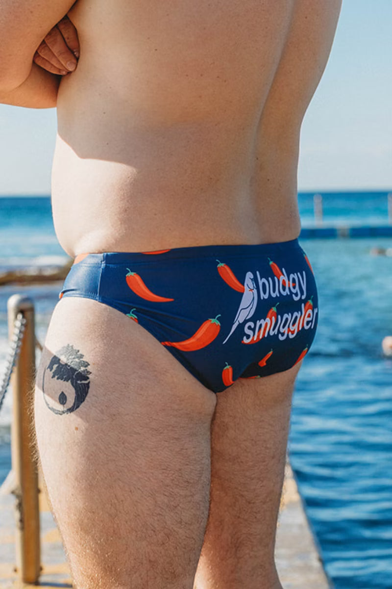 A close-up lifestyle shot of a person from behind wearing navy blue swim briefs with a red chili pepper pattern and the text 'budgy smuggler' printed on the side, set against a background of blue water and sky.