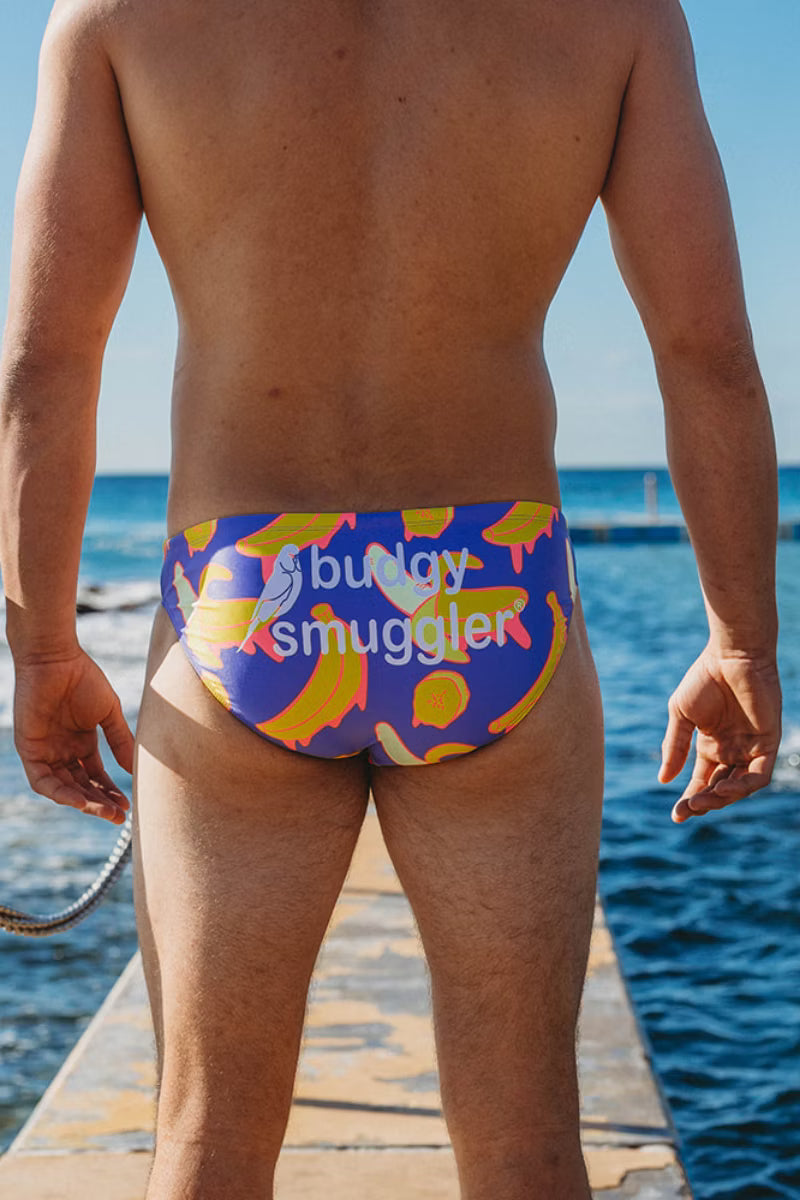 A man viewed from behind wearing colorful swim briefs with a blue, yellow, and orange abstract pattern and the text 'bully smuggler' printed across the back, standing on a dock with the ocean in the background.