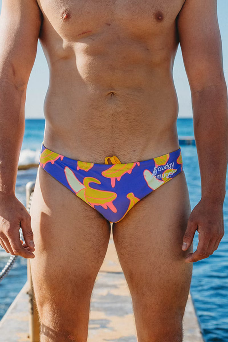A man wearing colorful swim briefs with a banana print, standing on a wooden dock with the ocean in the background.