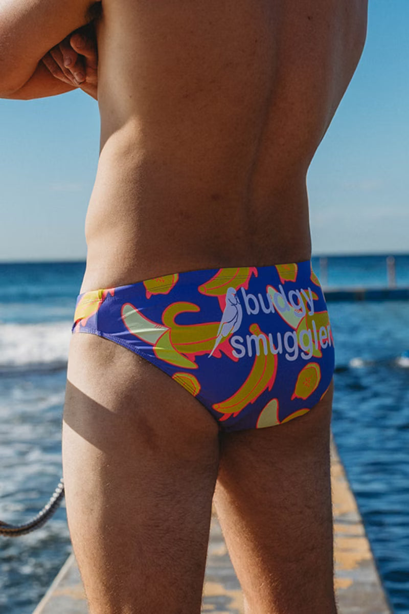A man wearing colorful swim briefs with a banana and bird pattern, standing on a wooden dock with the ocean and blue sky in the background.
