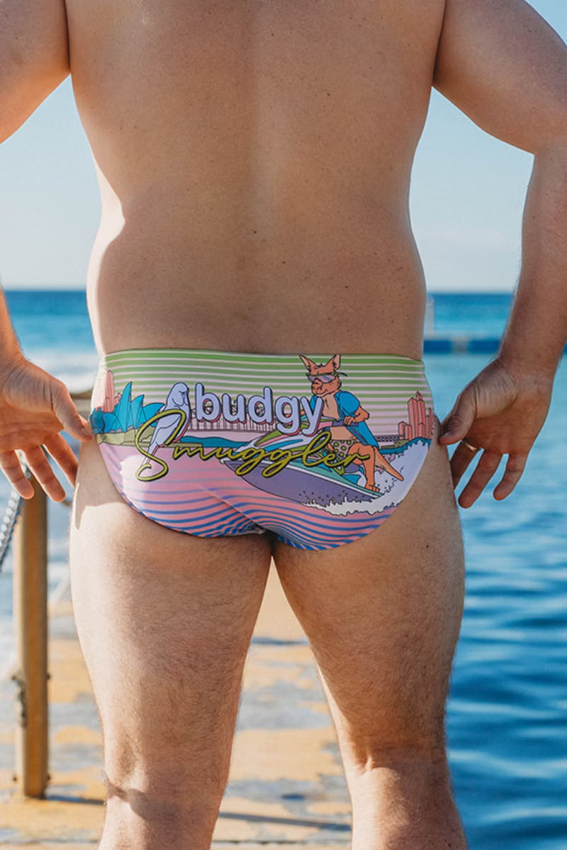A man standing on a beach wearing low-rise swim briefs with a colorful retro graphic featuring the text 'Cibudy Smully Stump' and a cartoon character.