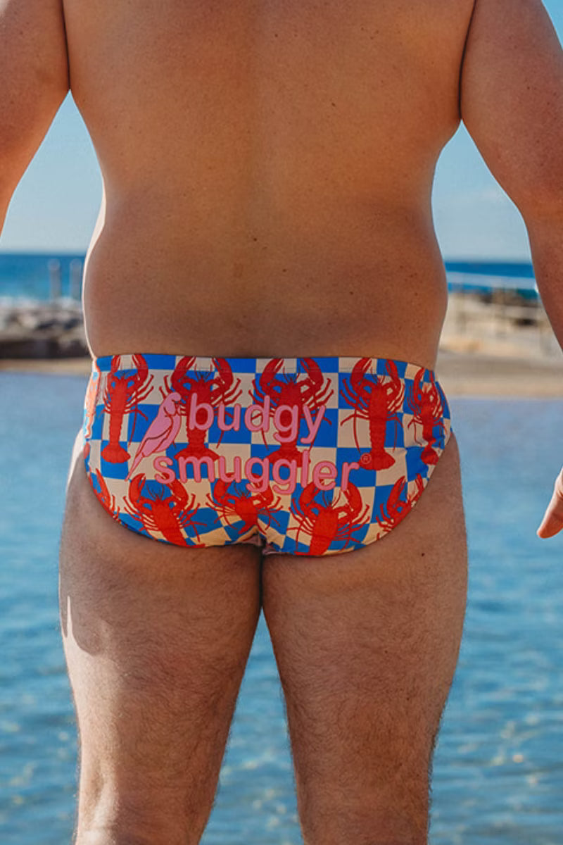 Rear view of a person standing at the beach wearing bikini bottoms with a lobster and flag pattern in red, blue, and white, with the text 'ribuddy swimwear' printed across the front.