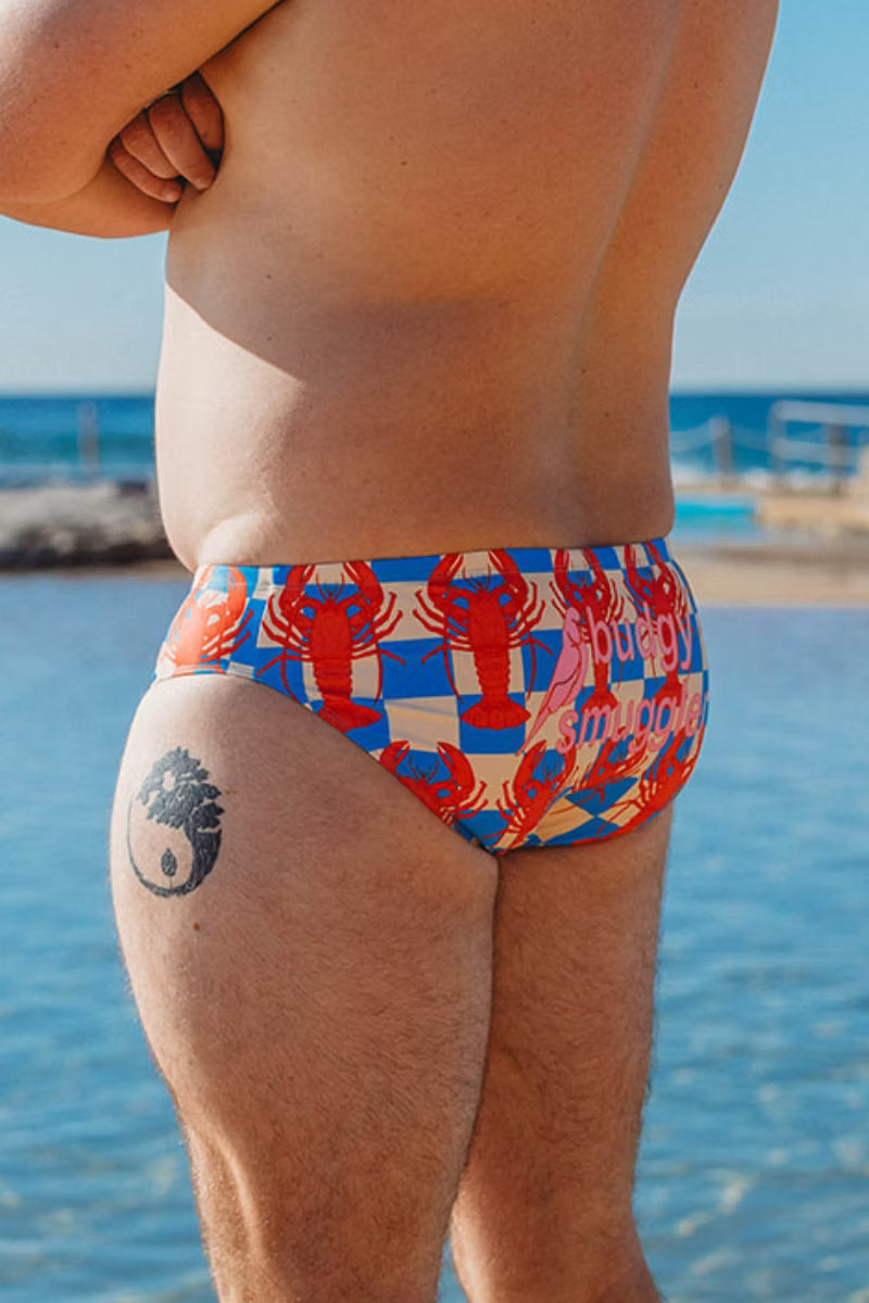 A man wearing swim briefs with a colorful lobster and checkered pattern, photographed from behind at a beach with blue water and sky in the background.