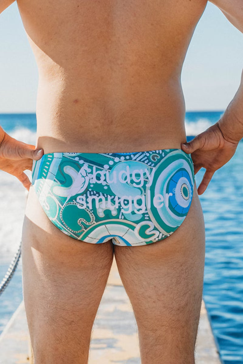 A man wearing swim briefs with a blue, teal, and white abstract pattern and the text 'Holiday Smuggler' on the back, standing on a dock with the ocean in the background.