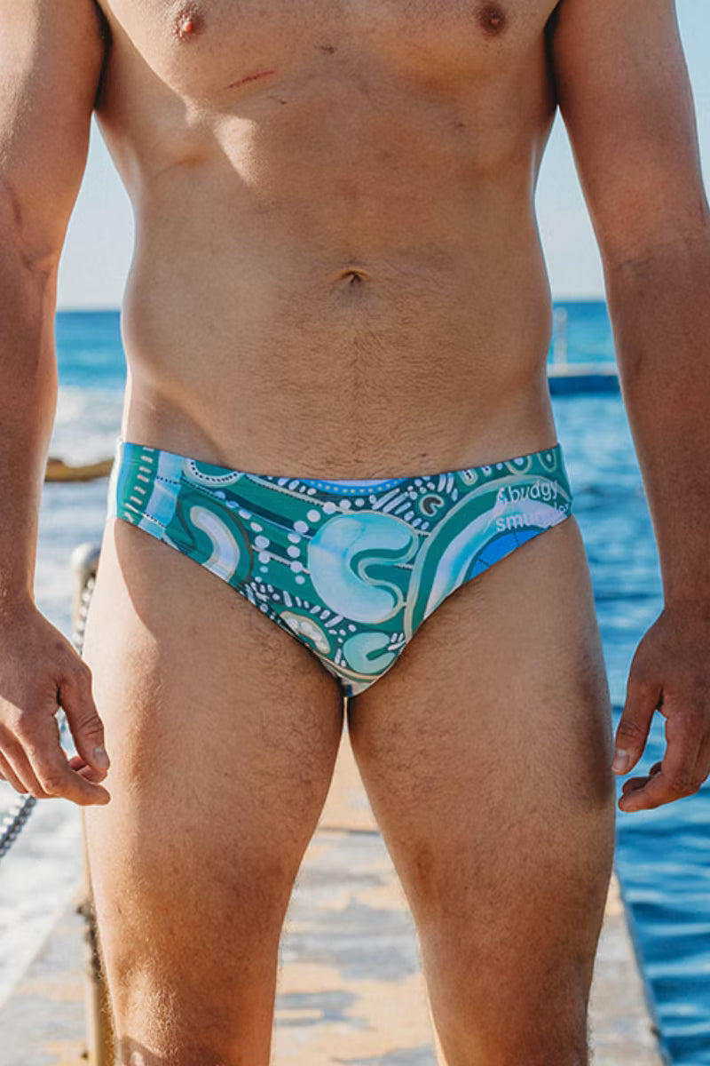 A close-up of a man's lower torso wearing swim briefs with a blue, teal, and white abstract pattern, standing on a sandy beach with the ocean in the background.
