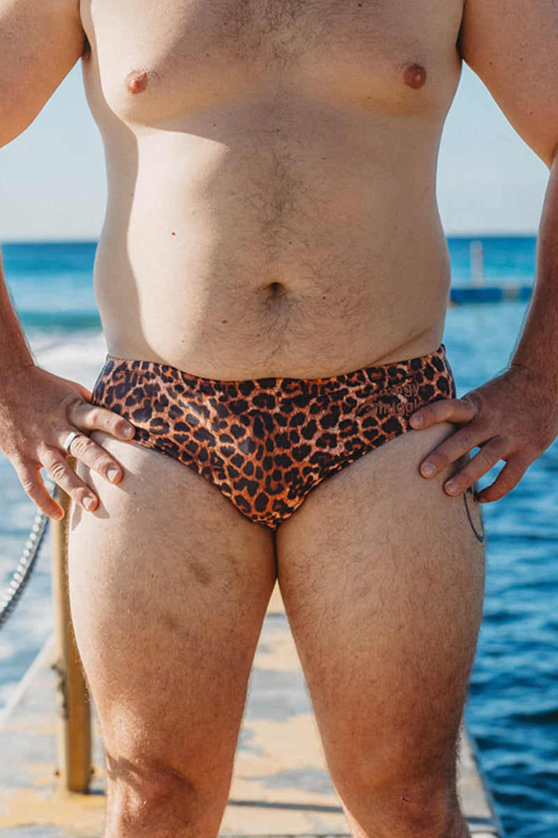 A close-up lifestyle shot of a man wearing leopard print swim briefs on a sunny day by the ocean.