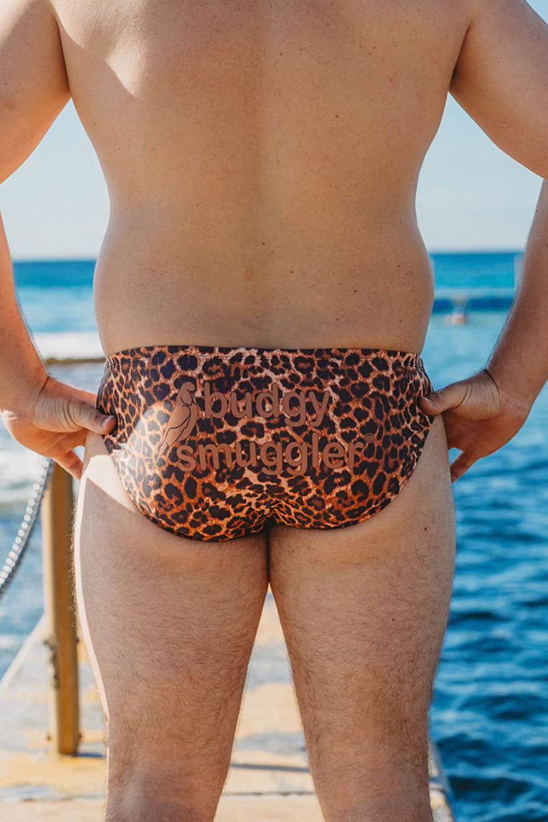 Rear view of a person wearing leopard-print swim briefs with the words 'surrounded by angels' printed across the front, standing outdoors near water.