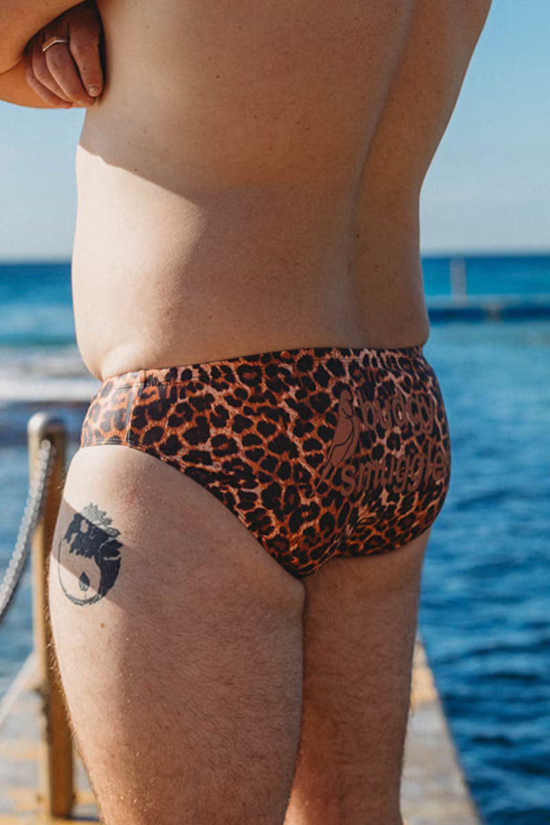 A close-up back view of a person wearing leopard-print swim briefs, standing outdoors near the ocean with blue water and sky in the background.
