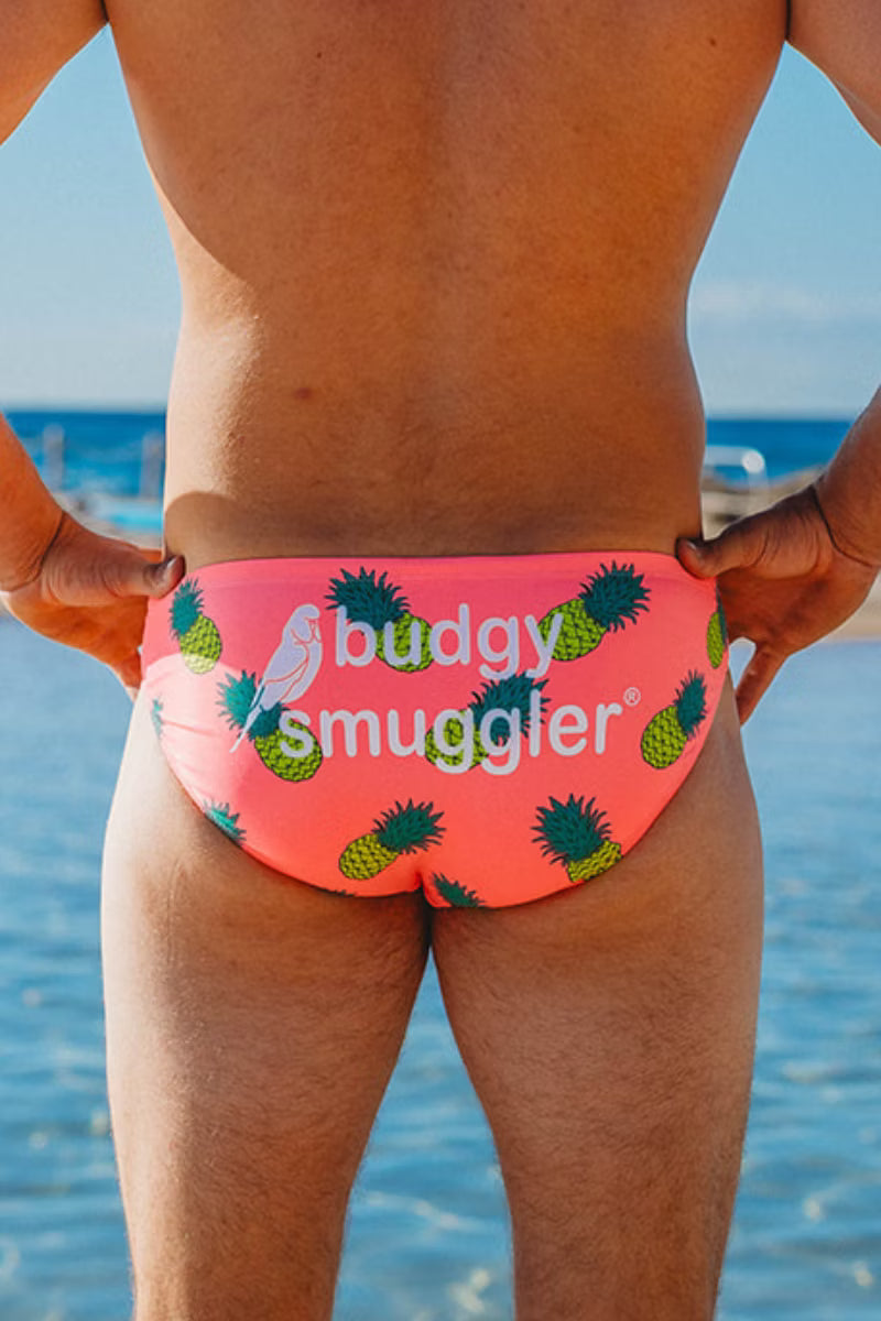 Rear view of a person wearing bright pink swim briefs with a green pineapple print and the text 'buddy smuggler' on the front, standing outdoors near water.