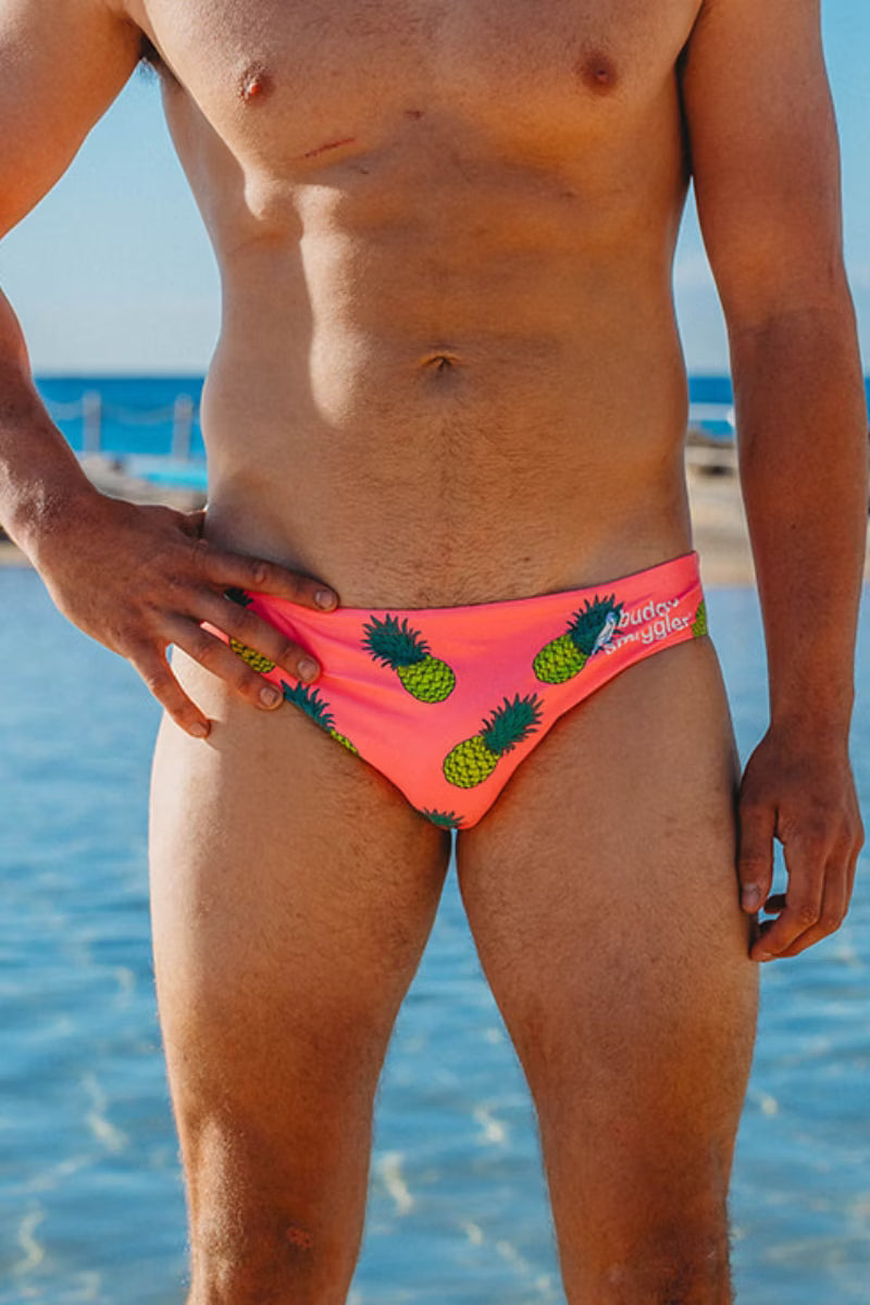 A close-up of a man wearing bright pink swim briefs with a green pineapple print, standing in shallow blue ocean water.