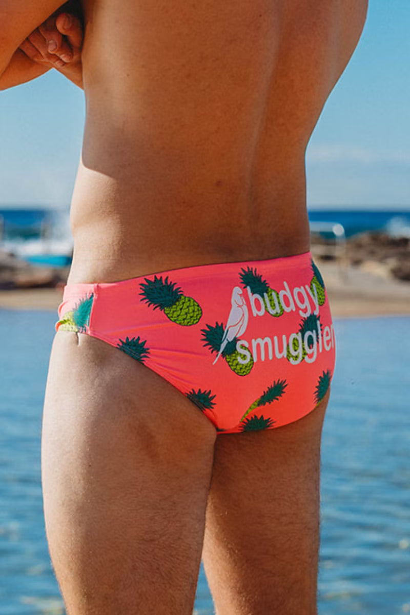 A close-up back view of a person wearing bright pink bikini bottoms with a pineapple print and the words 'buddy smuggler' on the back, standing by the ocean.