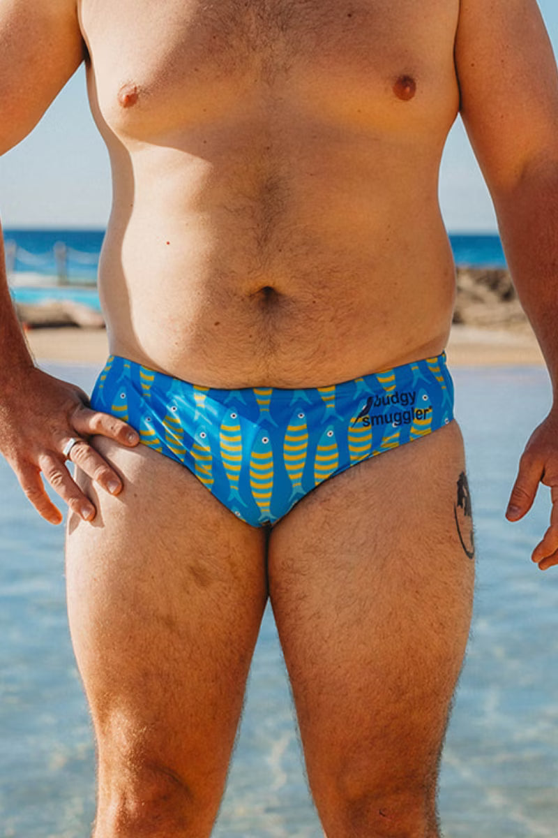 A close-up of a man's lower torso and thighs at the beach, wearing blue swim briefs with a yellow fish pattern and the brand name 'Buddy Smugger' printed on the front.