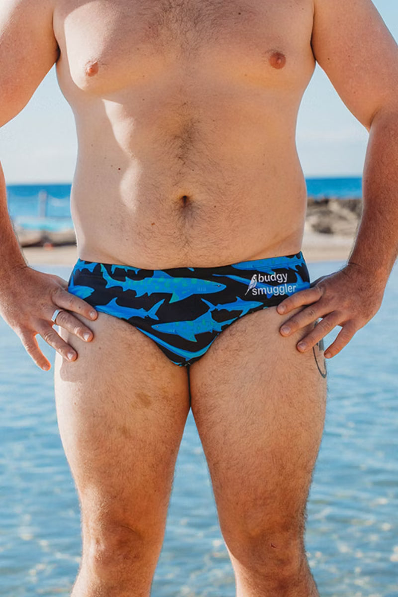 A man wearing blue and black shark-patterned swim briefs with the brand name 'buddy smuggler' printed on the side, standing on a beach with water in the background.