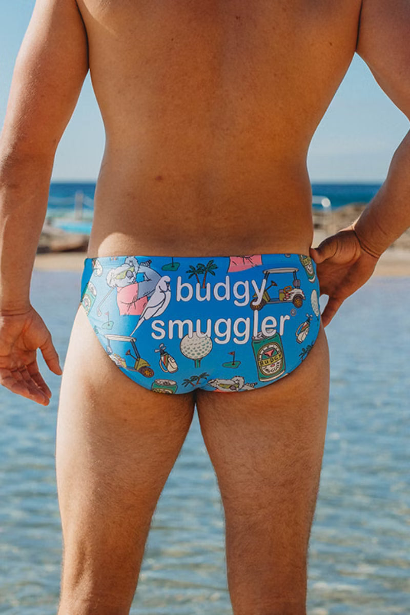 A man standing on a beach wearing blue swim briefs with a colorful cartoon print and the words 'budgy smuggler' across the front.